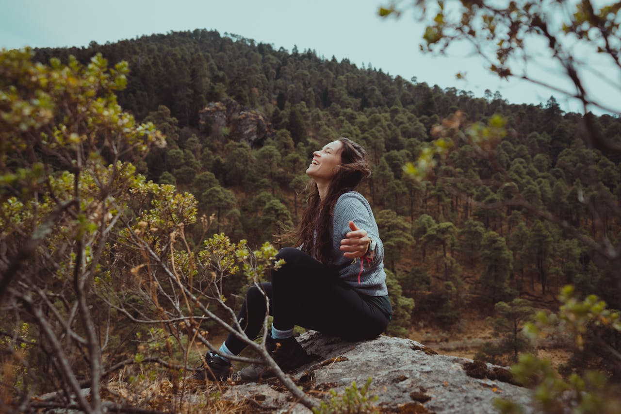 A happy woman sitting on a boulder with a mountain and trees in the background